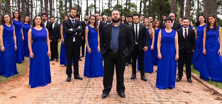 #OSUAcessível Fotografia com o Coro Contemporâneo. Ao centro e a frente o Regente Angelo Fernandes veste traje social preto. Ele tem pele branca e usa barba escura. Atrás dele está todo o coro. Os homens vestem terno preto, camisa social branca e gravata preta e as mulheres vestido longo azul royal. Todos estão sérios.
